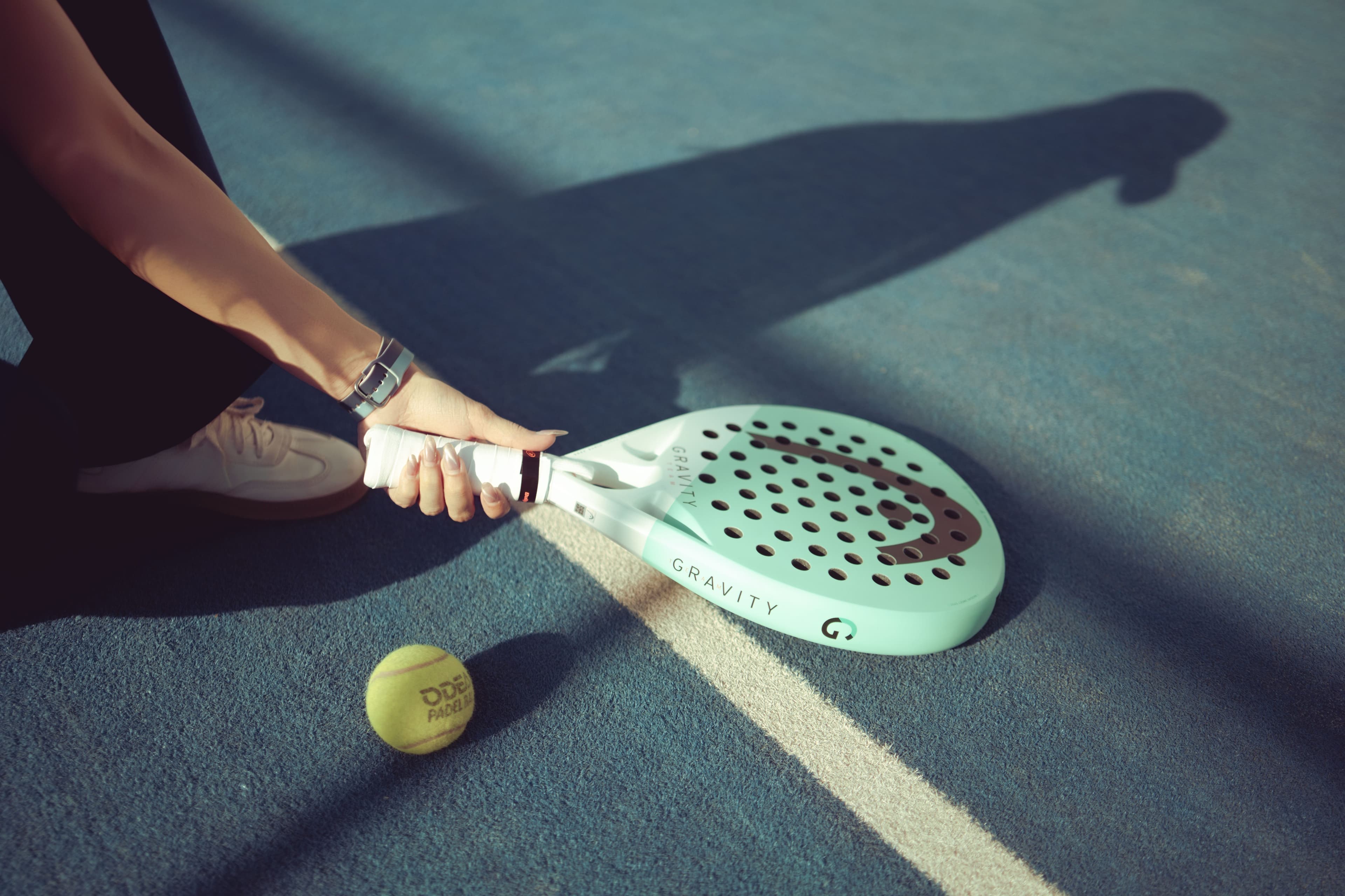 A padel player kneeling on a blue court at golden hour, gripping a paddle beside a ball